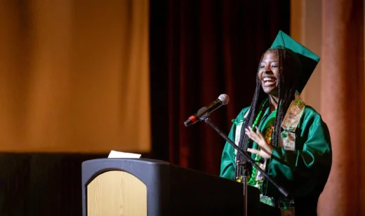 A smiling Black graduate in a green cap and gown speaks excitedly at a podium. TITLE: