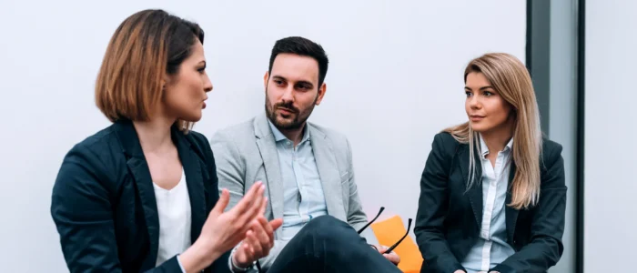 Three business professionals are engaged in a discussion, with one woman speaking and gesturing.