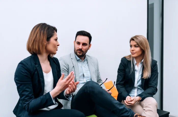 Three business professionals are engaged in a discussion, with one woman speaking and gesturing.