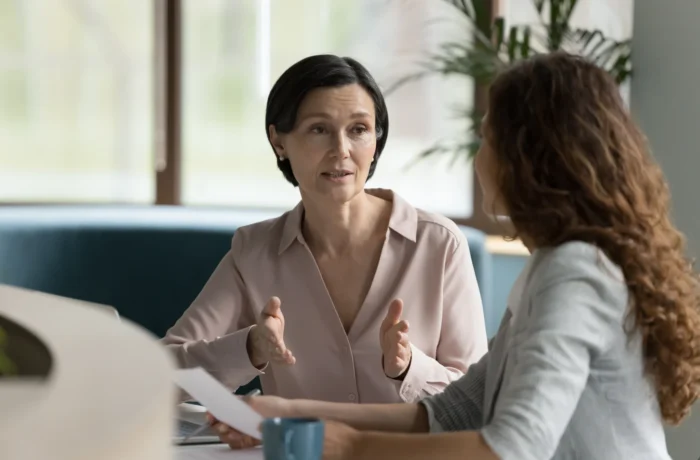 A mature woman gestures while earnestly speaking to a younger colleague in an office setting. TITLE: