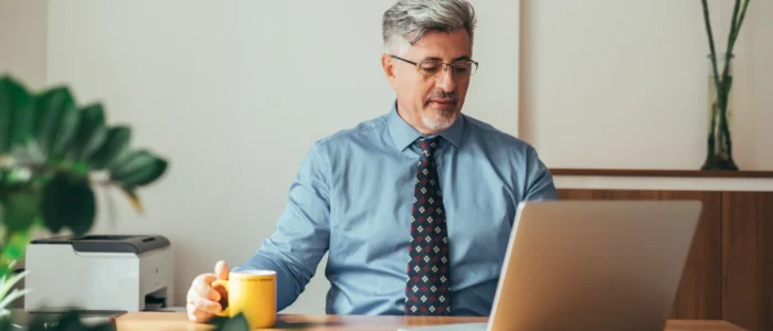 A professional man in a blue shirt and tie works on his laptop at a desk, holding a