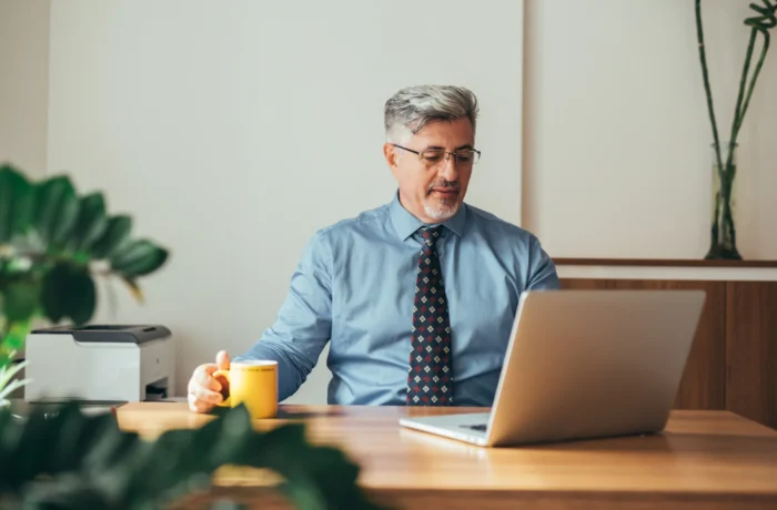 A professional man in a blue shirt and tie works on his laptop at a desk, holding a